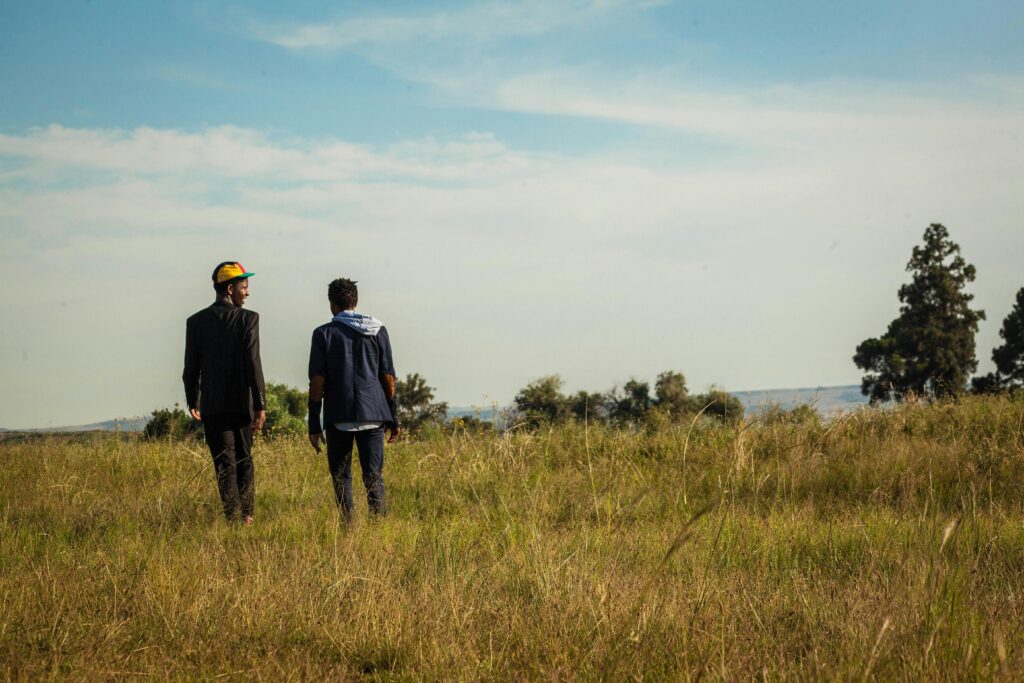 Back view of unrecognizable males strolling in rural meadow with dry grass and green trees and talking to each other